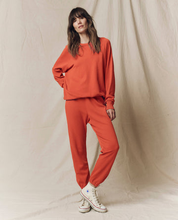 A person stands on a light fabric backdrop, wearing THE GREAT.'s The College Sweatshirt in Solid Desert Clay from the SU25 SEASONAL KNITS collection, paired with matching pants and white high-top sneakers.