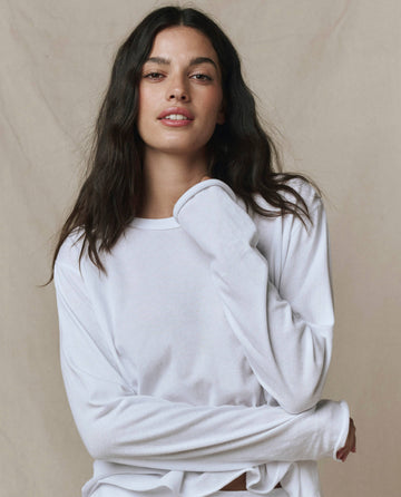 A person with long brown hair wears THE GREAT. CORE KNITS The Long Sleeve Crop Tee in True White, featuring a raw hem, standing before a beige background and gazing neutrally at the camera.