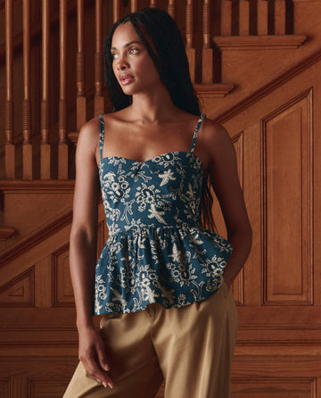 A woman stands indoors before a wooden staircase, wearing THE GREAT. SP26 D1's The Verse Top in Indigo Bandana—a trendy blouse that pairs perfectly with beige pants as she gazes to the side.