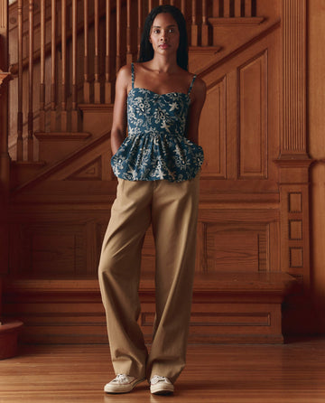 A woman stands indoors by wooden stairs, wearing THE GREAT. SP26 D1 The Verse Top in Indigo Bandana, tan wide-leg pants, and white sneakers—a stylish choice for women's fashion.