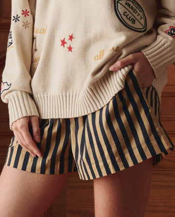 A person stands indoors wearing a cream sweater with embroidered patches and THE GREAT. SP26 D1 The Boxer Short in Khaki/Black Club Stripe.