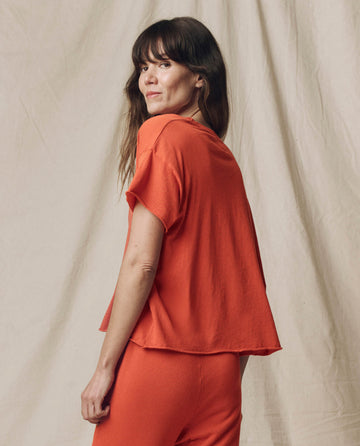 A woman with long brown hair wears THE GREAT. SU25 SEASONAL KNITS’ The Crop Tee. Solid in Desert Clay, paired with matching pants, standing against a neutral fabric backdrop—a chic update on casual women’s style.