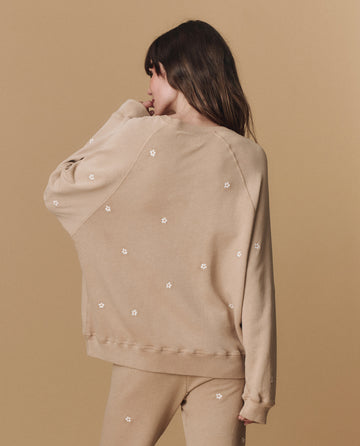 A person faces away from the camera wearing THE GREAT. PF25 EMB KNITS' The Slouch Sweatshirt—khaki with daisy embroidery—paired with matching pants, set against a beige background.