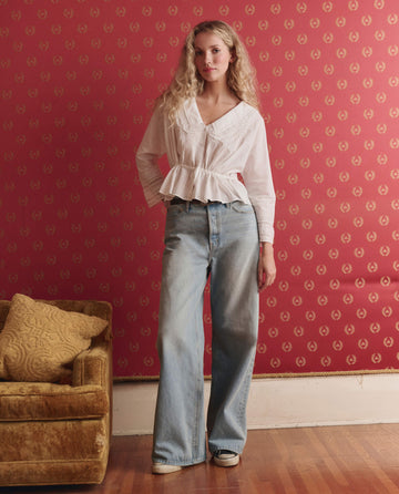 A woman stands indoors against a red patterned wall, wearing a white blouse and THE GREAT. F25 D1 The Slouch Wide Leg Jean in Tidal Wash, with one hand in her pocket. A gold couch with pillows is beside her.