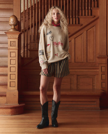 A person with long curly blonde hair stands indoors by a wooden staircase, wearing a beige graphic sweater, black cowboy boots, and THE GREAT. SP26 D1 The Boxer Short. -- Khaki/Black Club Stripe.