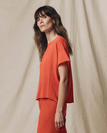 A woman with long brown hair models THE GREAT. SU25 SEASONAL KNITS The Crop Tee. Solid in Desert Clay, paired with matching pants against a beige fabric backdrop, exemplifying easy women's fashion.