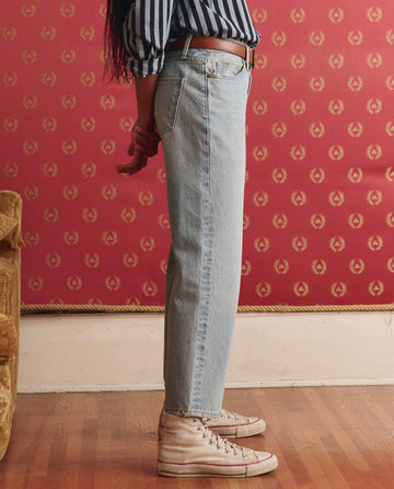 A person stands sideways indoors in THE GREAT. SP26 D1 The Wayne Jean -- Tidal Wash, a striped shirt, and white high-top sneakers against a red patterned wall and wooden floor.