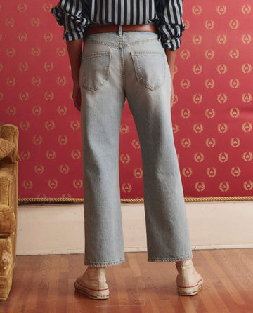 A person stands indoors on a wooden floor, facing away, wearing THE GREAT. SP26 D1 The Wayne Jean in Tidal Wash, a striped shirt, and white shoes in front of a red patterned wall.