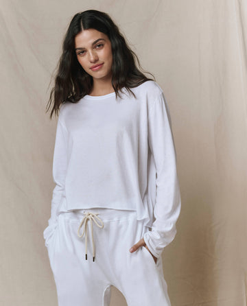A woman stands with hands in pockets against a neutral background, wearing THE GREAT. CORE KNITS’ The Long Sleeve Crop Tee in True White and drawstring pants.