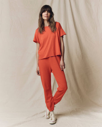 A person stands against a beige fabric backdrop, modeling THE GREAT. SU25 SEASONAL KNITS "The Crop Tee. Solid -- Desert Clay" paired with matching pants and white sneakers for a stylish casual look.