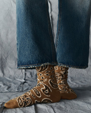 A person stands on a gray textured surface, wearing blue jeans and THE GREAT. H25 COLLECTION's The Bandana Sock in Camel, featuring a paisley pattern.