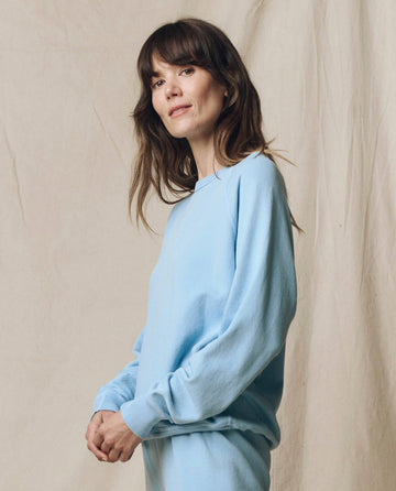 A person with long brown hair wears THE GREAT. SU25 SEASONAL KNITS "The College Sweatshirt. Solid -- Vista Blue" and stands before a beige fabric backdrop, looking at the camera.