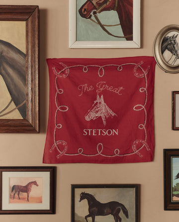 The Stetson Western Bandana in red by THE GREAT. STETSON SP26, featuring a horse head design and "The Great Stetson" text, is displayed on a wall surrounded by framed horse artworks.