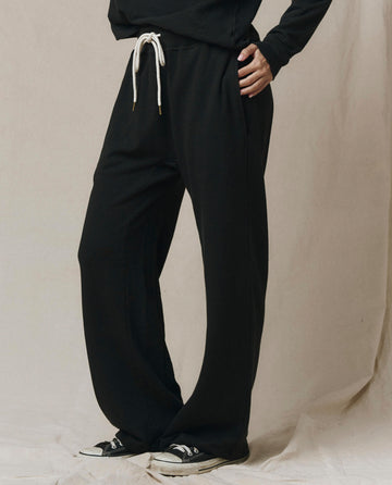 A person stands with hand on hip, wearing SP26 New Knits' The Horseshoe Sweatpant in Solid -- Almost Black with a white drawstring and black sneakers, set against a neutral background.