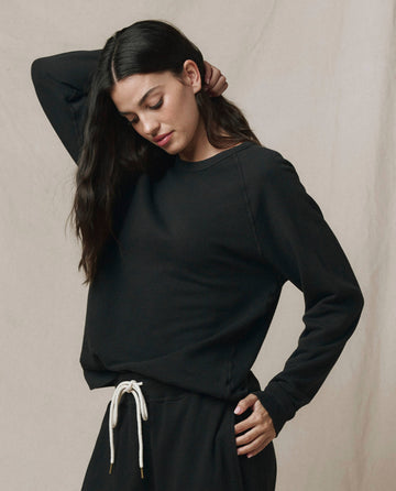 A woman wearing SP26 New Knits' The Rookie Sweatshirt in Solid -- Almost Black and drawstring pants stands against a neutral backdrop, one hand resting on the back of her head.