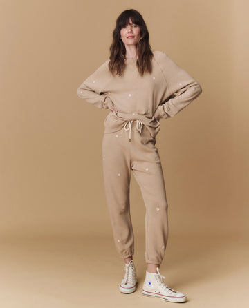 A woman wears THE GREAT. PF25 EMB KNITS’ The Stadium Sweatpant. Embroidered in khaki with daisy embroidery, paired with a matching sweatshirt and white high-top sneakers, standing against a beige backdrop.