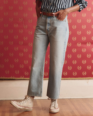A person wears THE GREAT. SP26 D1 The Wayne Jean in Tidal Wash—cropped, light blue 100% cotton denim—paired with a striped shirt and white high-top sneakers, standing on a wooden floor before a red patterned wall.