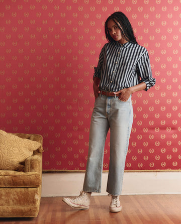 A woman stands indoors by a red patterned wall, wearing a striped shirt, THE GREAT. SP26 D1 The Wayne Jean in Tidal Wash, and white sneakers next to a gold sofa with a yellow pillow.