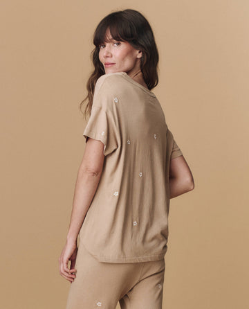 A woman with long brown hair models THE GREAT. PF25 EMB KNITS "The Boxy Crew. Embroidered" tee in khaki with daisy embroidery, paired with matching pants, standing against a beige background and looking over her shoulder.