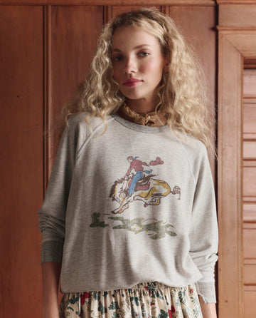 A woman with long curly blonde hair wears THE GREAT. SP26 D1 The College Sweatshirt w/ Rider Graphic in Soft Heather Grey and a floral skirt, standing before wooden paneling.