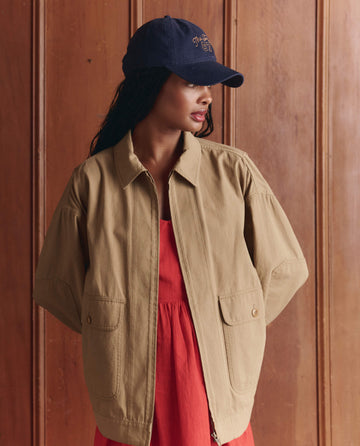 A woman in THE GREAT.'s The Folsom Jacket—Khaki (SP26 D1) over a red dress and navy baseball cap stands before a wood-paneled wall, gazing left.