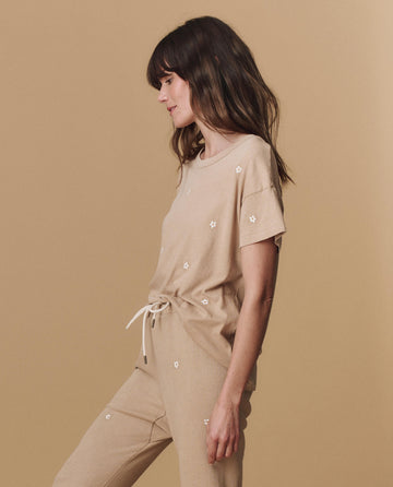 A woman poses in profile wearing THE GREAT. PF25 EMB KNITS "The Boxy Crew. Embroidered"—a khaki short-sleeve top with daisy embroidery—paired with matching pants, set against a plain beige background.