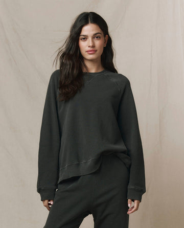 A woman with long dark hair wears THE GREAT. CORE KNITS The Slouch Sweatshirt in Solid Washed Black, paired with matching pants, standing against a plain light background.