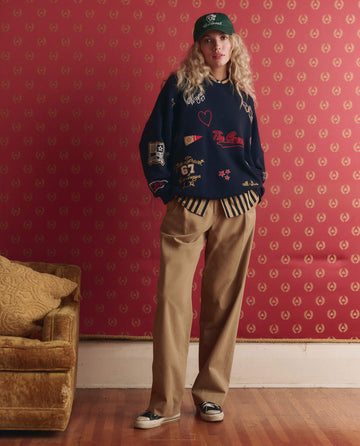 A person wearing a green cap, navy graphic sweater, THE GREAT. PS26 COLLECTORS "The Menswear Trouser" in khaki, and sneakers stands by a gold couch against red patterned wallpaper.