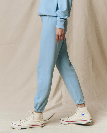 A person wears THE GREAT.'s The Stadium Sweatpant, Solid -- Vista Blue from the SU25 SEASONAL KNITS collection, paired with a matching sweatshirt and white high-top Converse sneakers, standing against a beige fabric backdrop.