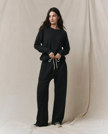 A woman stands before a beige fabric backdrop, wearing the SP26 New Knits "The Rookie Sweatshirt" in Solid -- Almost Black and matching black pants with white drawstrings.