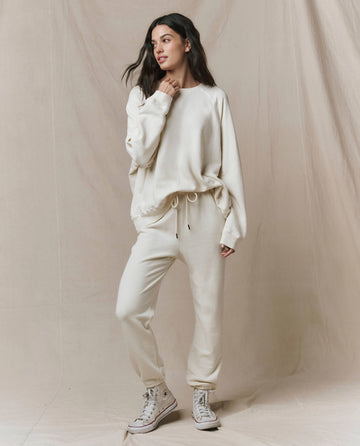 A woman stands against a beige backdrop wearing THE GREAT. CORE KNITS The Slouch Sweatshirt in Solid -- Washed White, matching drawstring sweatpants, and light-colored high-top sneakers.