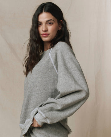 A woman with long dark hair wears THE GREAT. CORE KNITS The Slouch Sweatshirt in Varsity Grey, featuring fuller sleeves and a straight silhouette, standing before a beige fabric backdrop.