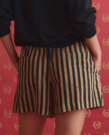 A person wearing THE GREAT. SP26 D1 The Boxer Short. in Khaki/Black Club Stripe stands facing a red wall with a gold wreath pattern, paired with a black top.