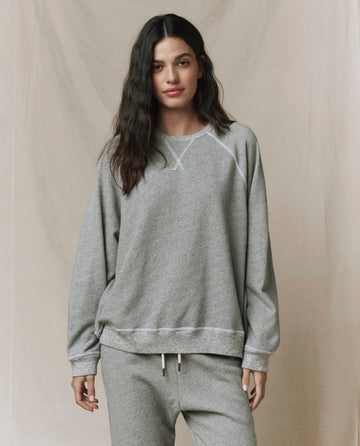 A person wears THE GREAT. CORE KNITS The Slouch Sweatshirt in Varsity Grey with matching straight-leg sweatpants, both featuring fuller sleeves, standing before a neutral background.