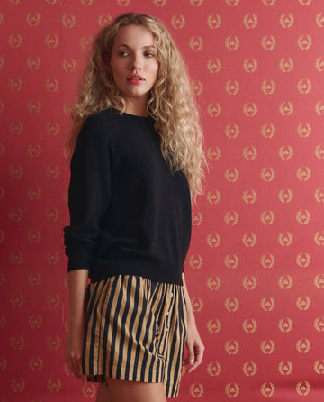 A woman with curly blonde hair wears a black sweater and THE GREAT. SP26 D1 The Boxer Short in Khaki/Black Club Stripe, standing before a red patterned wall.