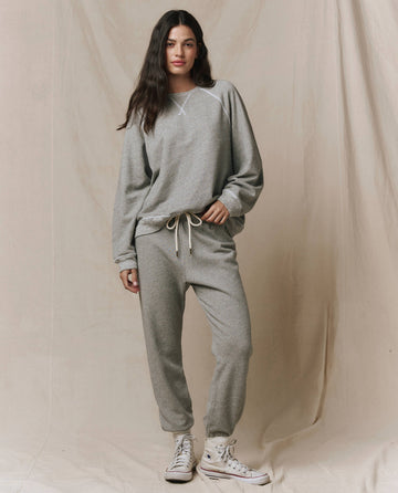 A woman stands before a beige backdrop wearing THE GREAT. CORE KNITS’ The Slouch Sweatshirt in Varsity Grey with fuller sleeves, paired with matching drawstring sweatpants and white high-top sneakers.