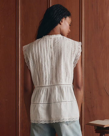 A person with long hair faces a wooden wall, wearing THE GREAT. SP26 D1 The Sonnet Top in True White and light blue jeans, featuring Victorian-inspired lace pintuck details.