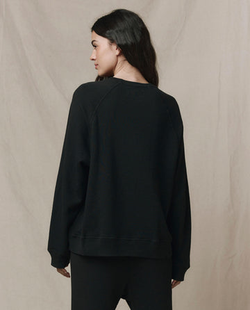 A person with long dark hair, facing away, wears THE GREAT. CORE KNITS The Slouch Sweatshirt in Solid -- Almost Black and black pants against a plain beige background.