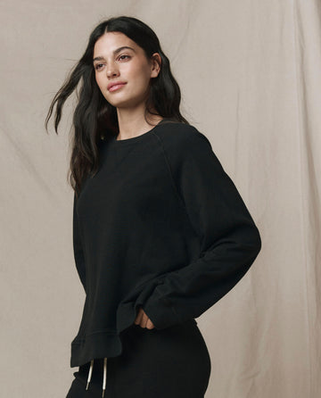 A woman with long dark hair wears THE GREAT. CORE KNITS' The Slouch Sweatshirt in Solid Almost Black, standing with one hand in her pocket against a neutral fabric backdrop.