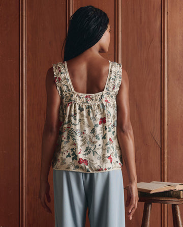 Facing a wooden wall, a person wears THE GREAT. SP26 D1’s The Victorian Pleat Tank -- Floating Butterfly and light blue pants, with an open book set on a nearby small table.