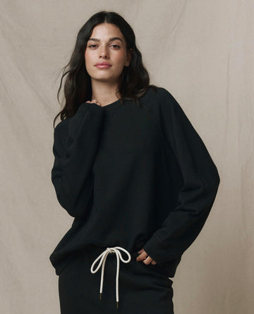A woman with long dark hair wears THE GREAT. CORE KNITS' Slouch Sweatshirt in Solid -- Almost Black, made of 100% cotton, paired with black pants featuring a white drawstring, standing against a neutral beige backdrop.