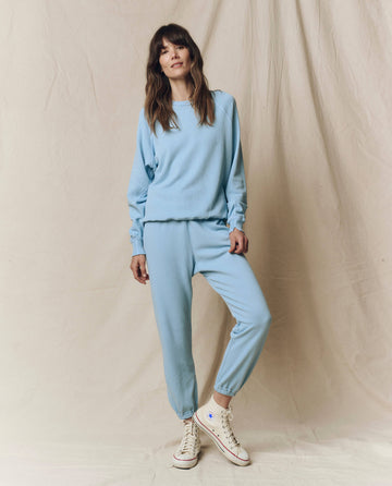 A woman models The Stadium Sweatpant. Solid in Vista Blue by THE GREAT. SU25 SEASONAL KNITS, standing with one foot forward against a beige fabric backdrop.