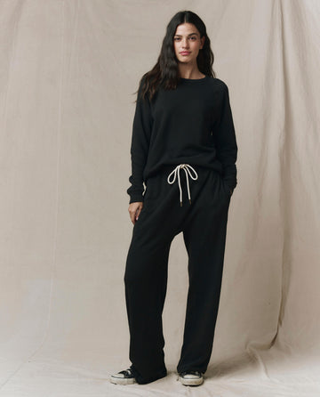A woman stands before a beige backdrop in SP26 New Knits’ The Horseshoe Sweatpant in Solid -- Almost Black, paired with a matching black long-sleeve sweatshirt and black-and-white sneakers.