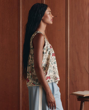 A woman with long braided hair in profile wears THE GREAT. SP26 D1's The Victorian Pleat Tank--Floating Butterfly with light blue pants, standing by a wooden wall and a small table holding an open book.