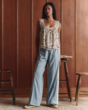 A woman stands indoors against a wood-paneled wall, wearing THE GREAT. SP26 D1 The Wide Leg Track Pant in Robin Blue with an elasticated waistband, a floral sleeveless top, and sneakers. Two wooden stools are nearby.