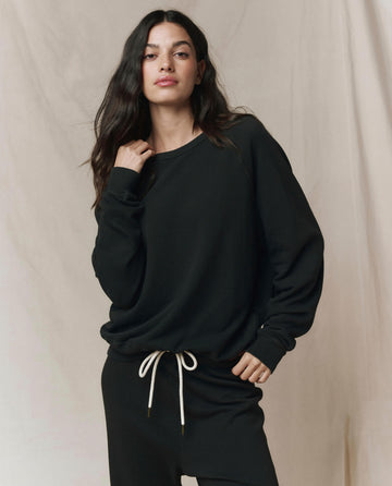 A person wearing THE GREAT. CORE KNITS The College Sweatshirt in Solid -- Almost Black and matching pants stands before a light-colored backdrop.