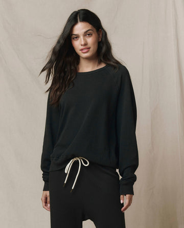 A person stands against a neutral background wearing THE GREAT. CORE KNITS The College Sweatshirt in Solid -- Almost Black, paired with black pants featuring a white drawstring.