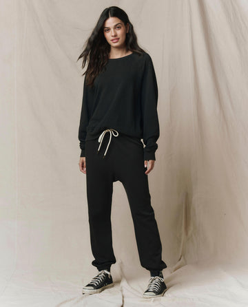 A person stands against a neutral fabric backdrop, wearing THE GREAT. CORE KNITS’ The College Sweatshirt. Solid -- Almost Black, paired with black drawstring pants and black high-top sneakers.