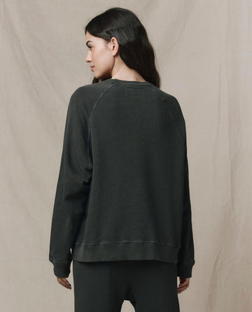 A woman stands facing away, wearing THE GREAT. CORE KNITS The Slouch Sweatshirt in Washed Black against a plain beige backdrop. Made in the USA.