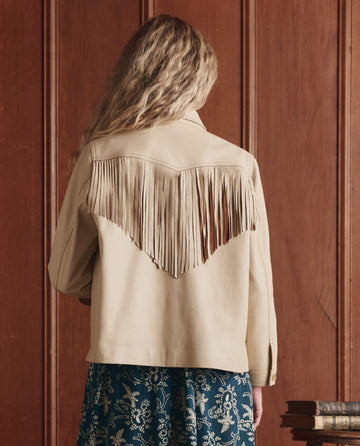 A person with wavy blonde hair wears THE GREAT. SP26 D1 The Leather Fringe Jacket in Toasted Almond, standing before a wooden wall.